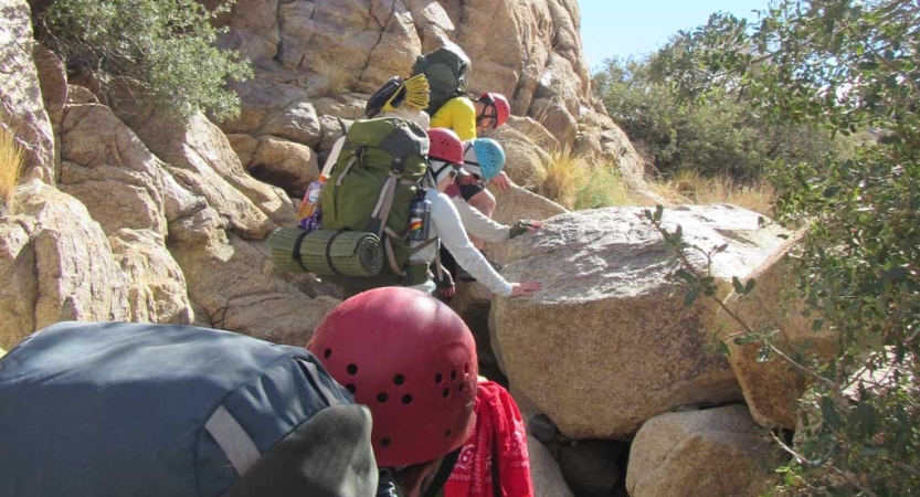 A group of people wearing backpacks and helmets hike across a rocky landscape, facing away from the camera. 